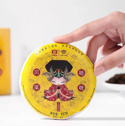 Hand holding a yellow dayi ripe pu erh tea cake packaging with intricate design on a white background